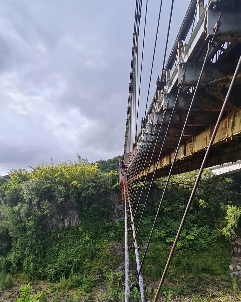 Sous le tablier du pont suspendu de Sainte-Rose, un cordiste progresse le long des câbles au-dessus de la ravine